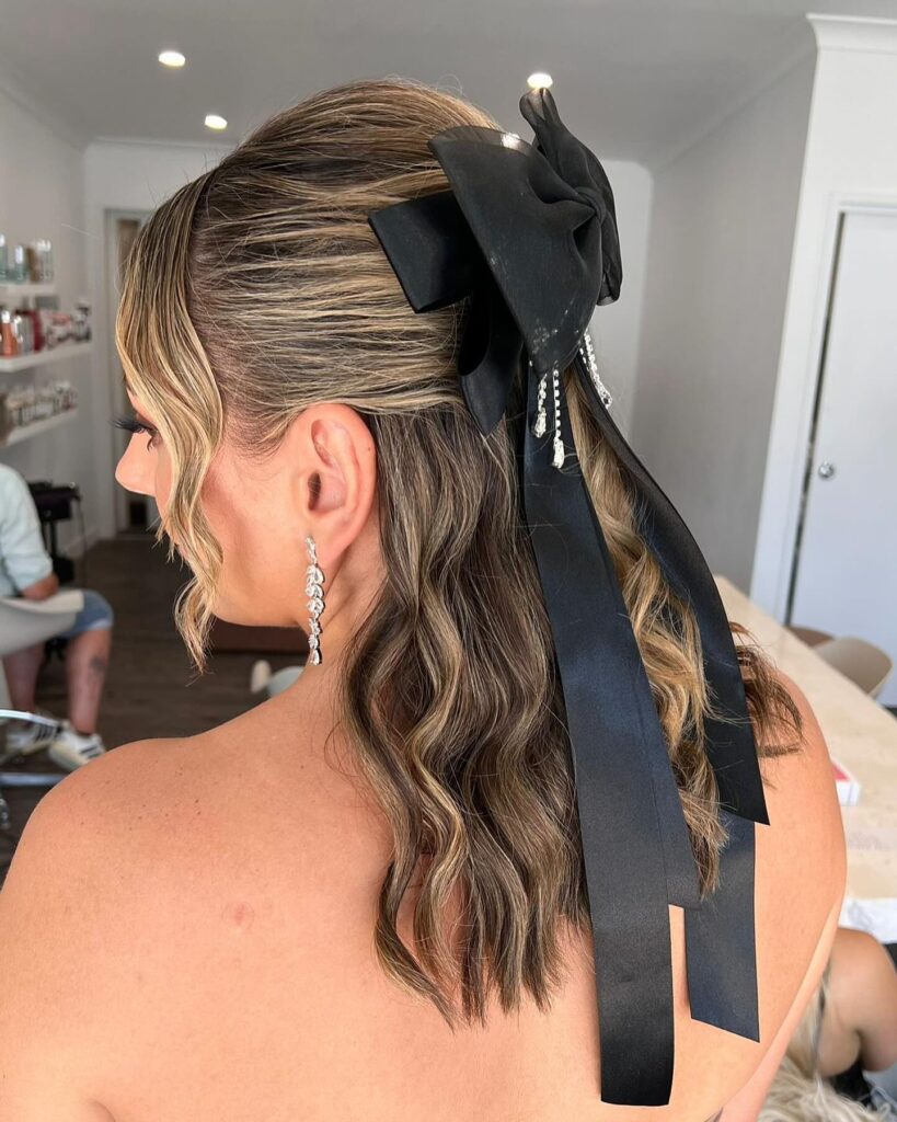 A woman viewed from the back wearing a half-up hairstyle with wavy brown hair and a large black bow adorned with dangling crystals.