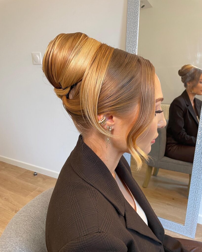 A side profile of a woman with a high, voluminous French twist updo in honey blonde tones with one wavy strand framing her face.
