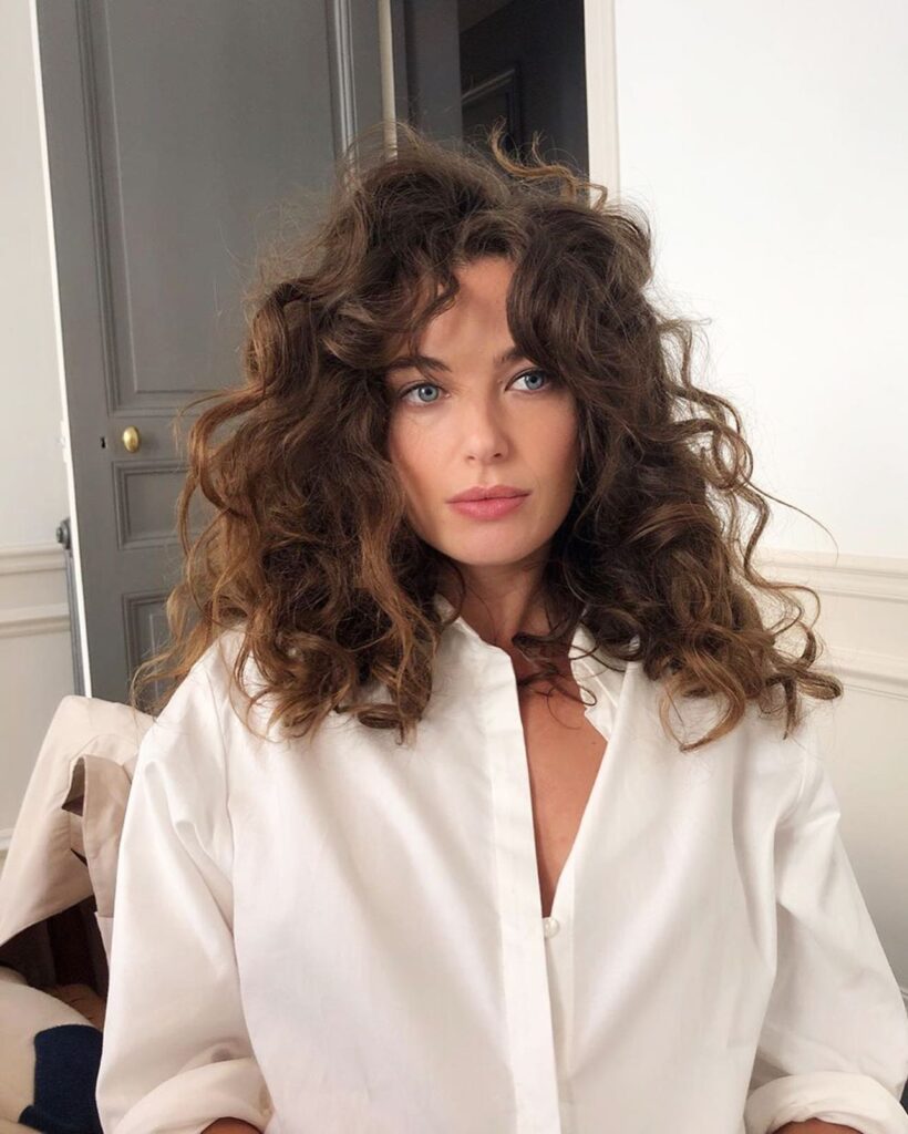 A woman with blue eyes and voluminous curly brown hair featuring wild texture and curly curtain bangs.