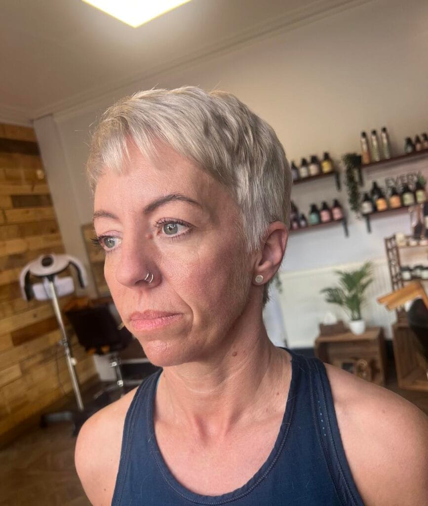 A woman showcasing a textured ash blonde micro pixie cut with short choppy bangs and tapered sides.