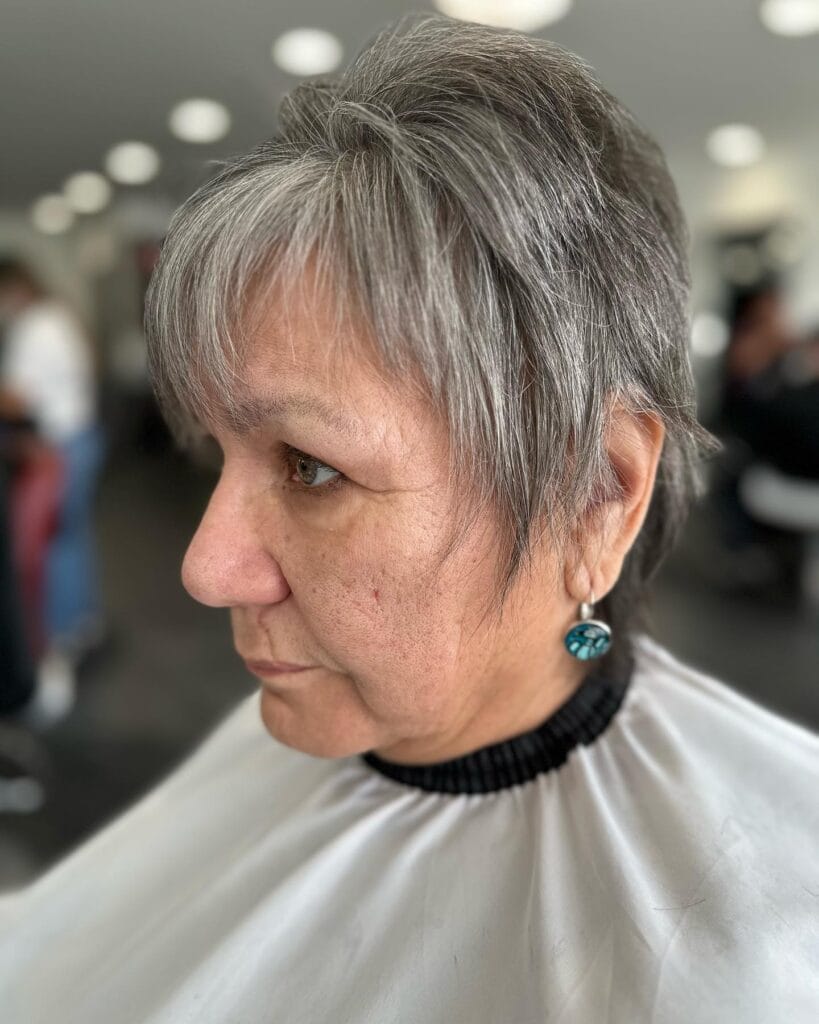 A side profile of a woman with a charcoal grey pixie cut featuring feathered layers and a wispy fringe.