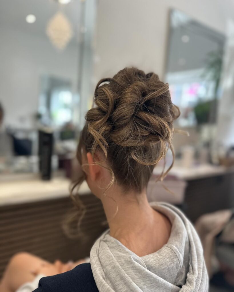 A loose high curly bun with soft volume and light brown highlighted strands.