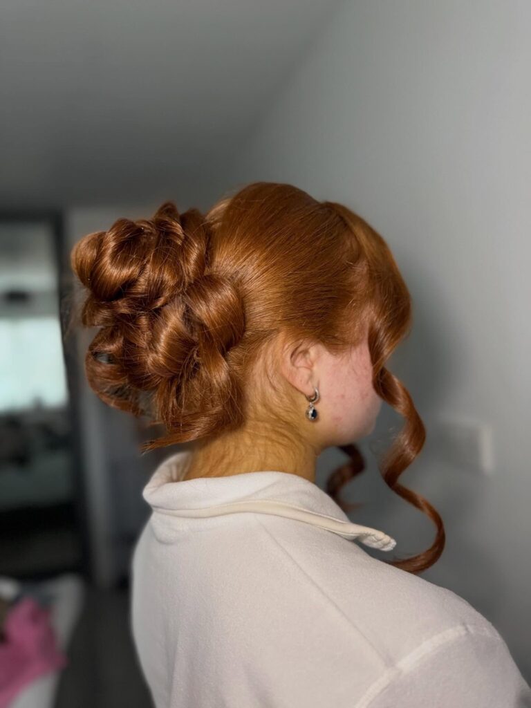 A glossy ginger curly bun with smooth roots and a loose curled strand framing the face.