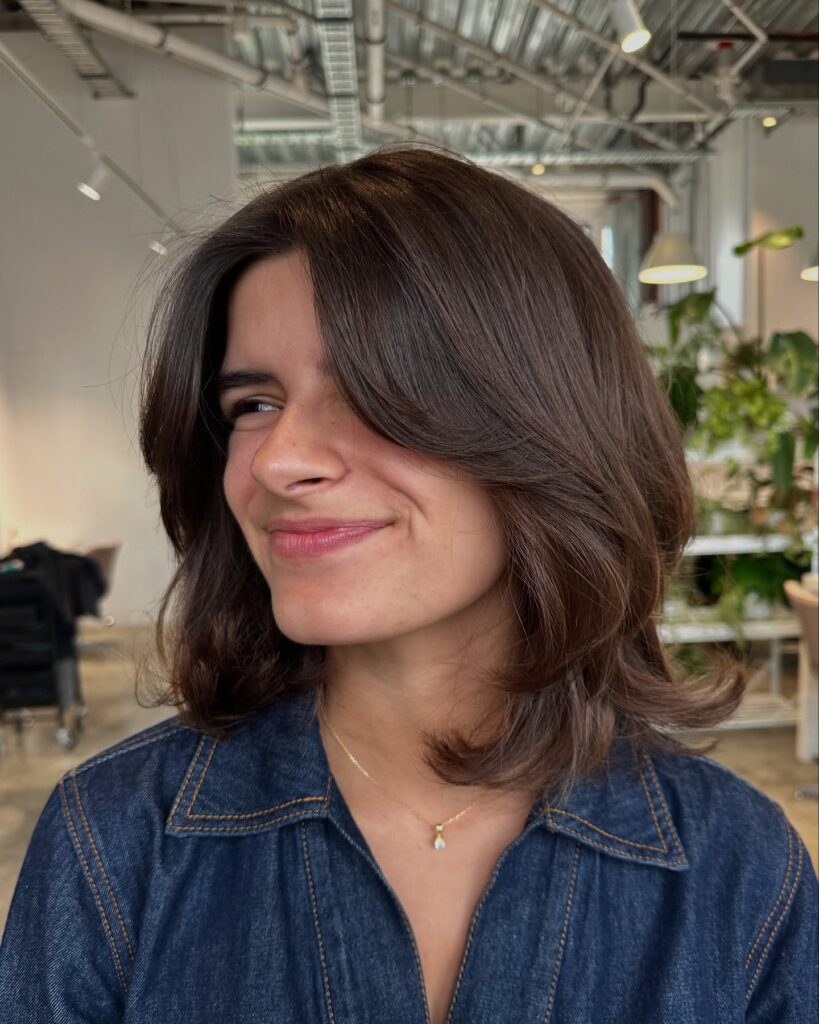 A brunette layered bob with soft waves and long side swept bangs creating a smooth face framing shape.