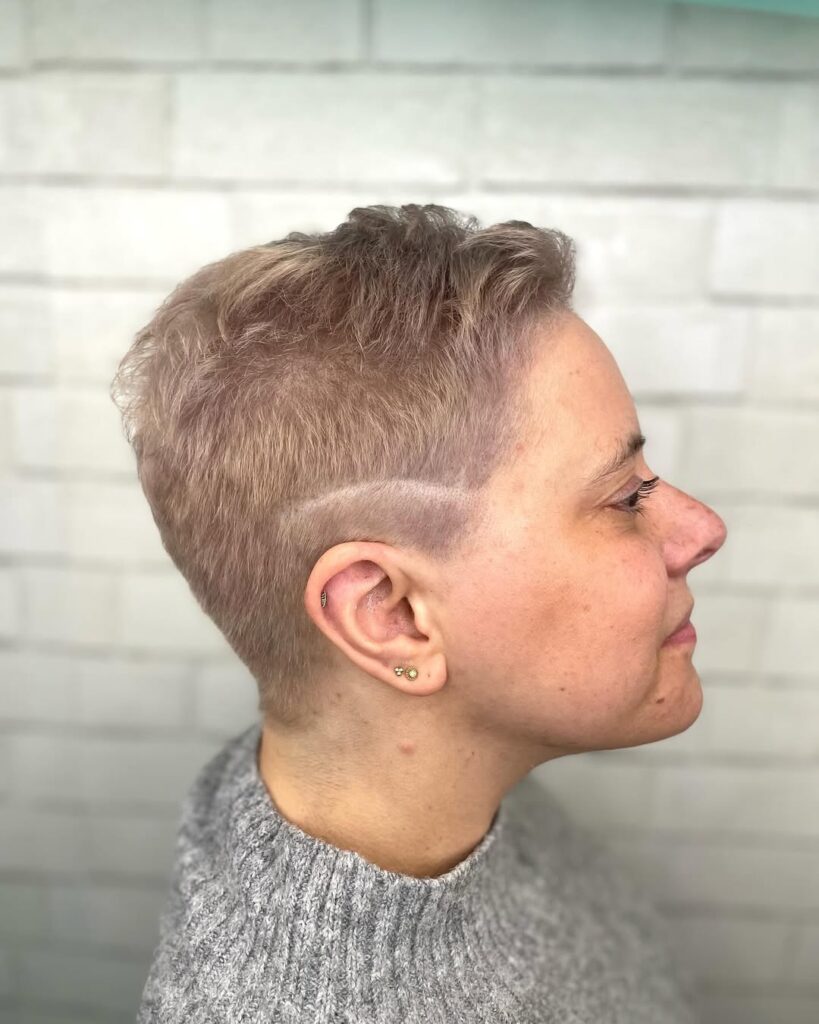 A woman with a textured blonde tomboy pixie haircut featuring shaved sides and a subtle line design with longer volume on top.