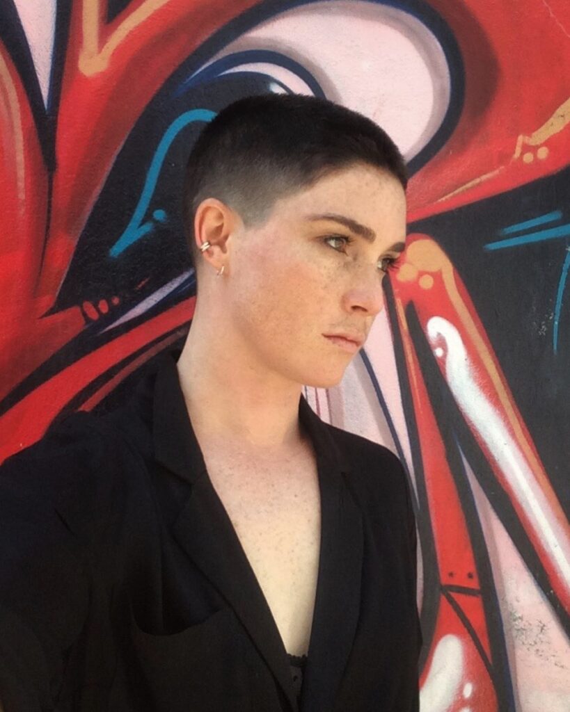 A woman with a very short dark buzz cut hairstyle standing in front of a colorful graffiti wall.