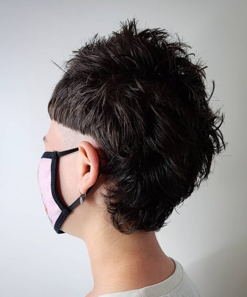 A woman with a tomboy mullet haircut featuring shaved sides and a longer textured back with messy dark layers.