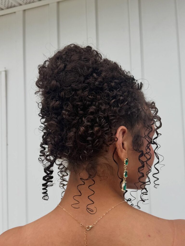 A high voluminous curly updo with tight coils and defined tendrils framing the face and neck.