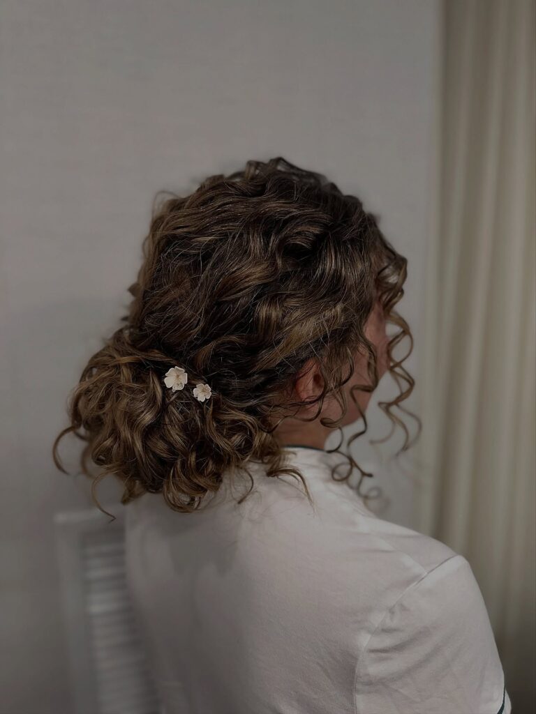 A soft low curly bun with loose waves and small decorative pins, with tendrils framing the face.