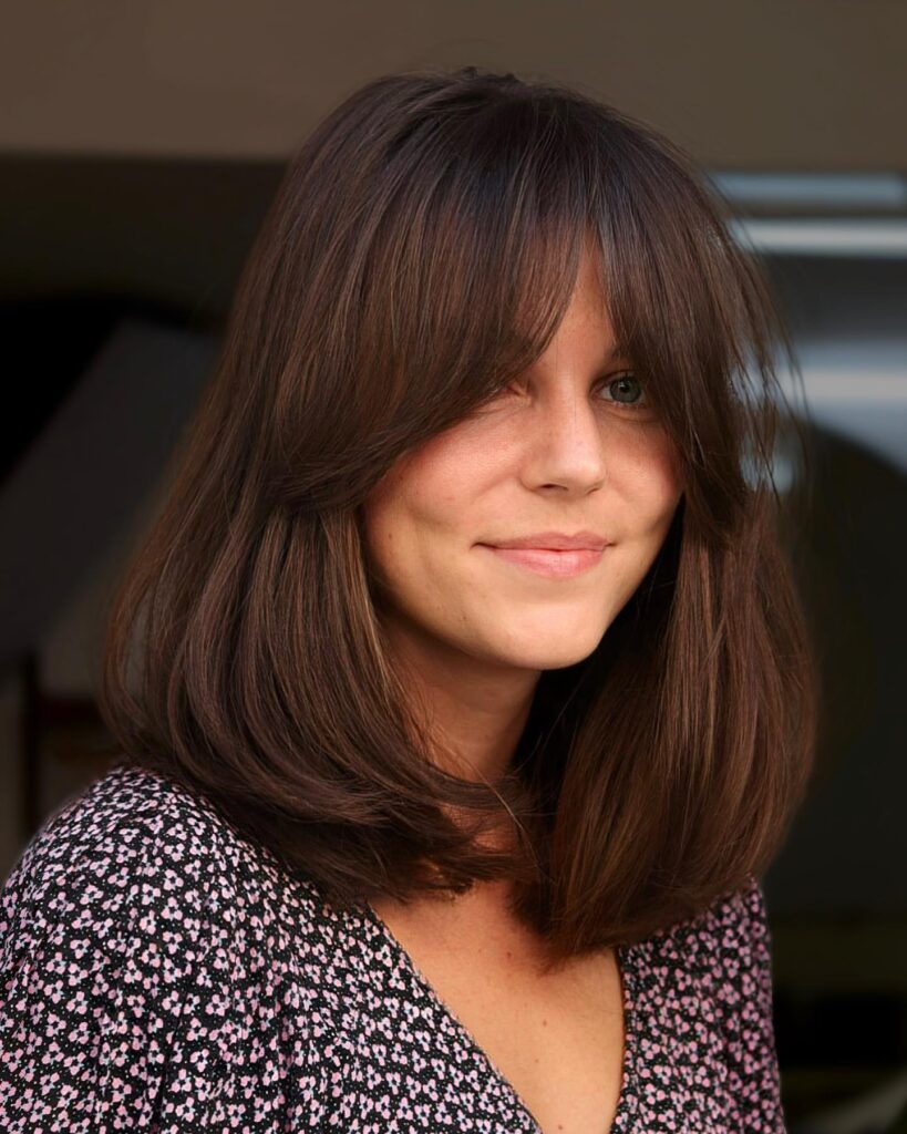 Mid length brown hairstyle with voluminous layers and full curtain bangs.