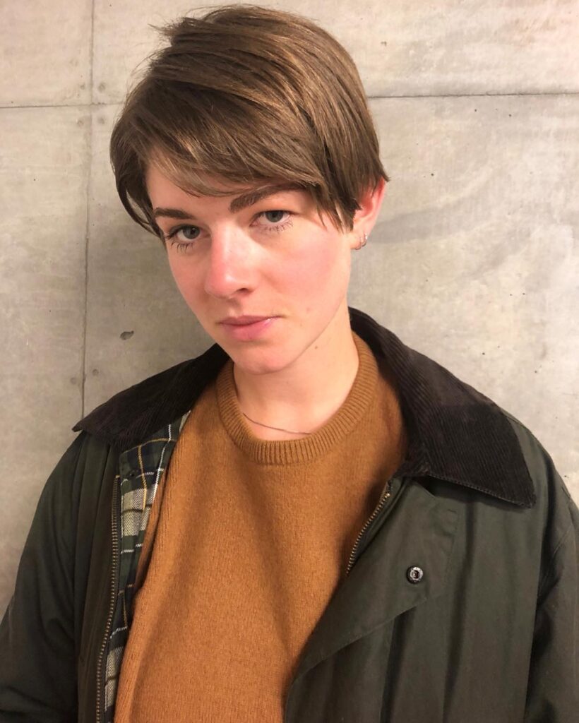A woman with a short tomboy haircut featuring a neat side part and softly tapered sides in light brown.