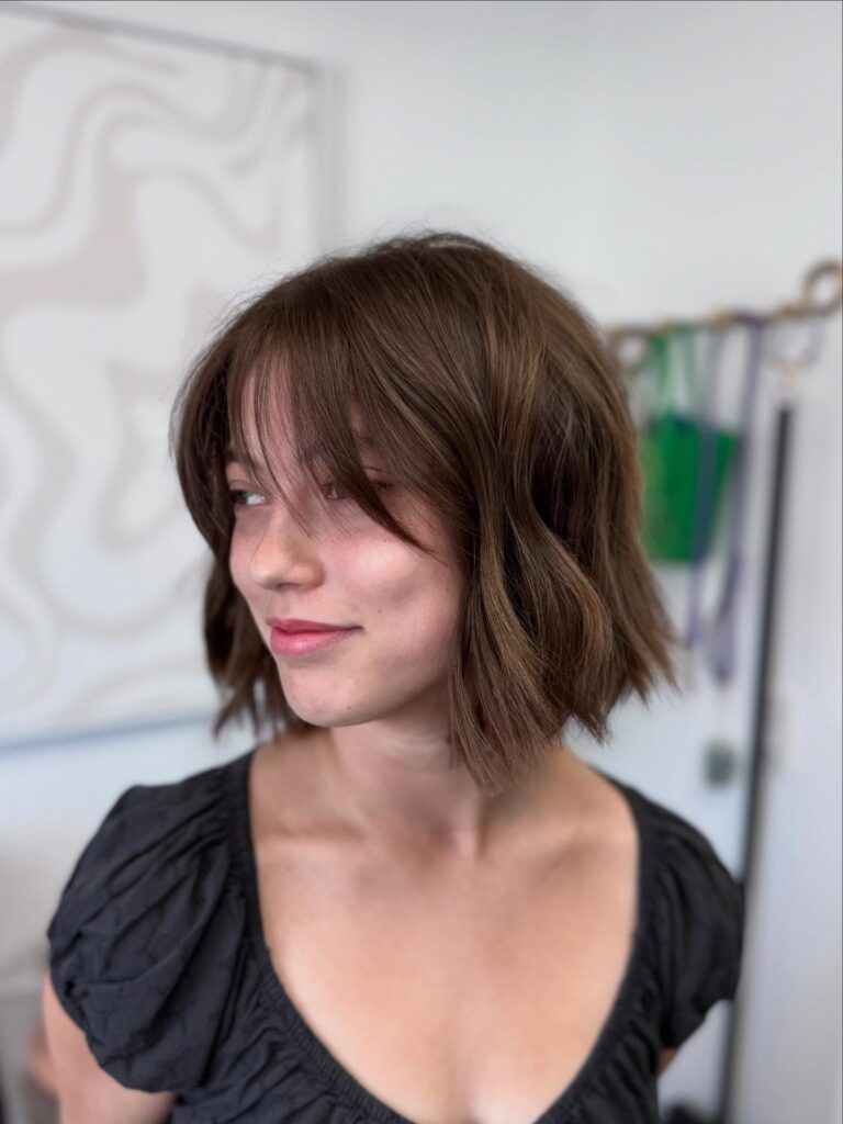 Chin length bob with soft layers and light wispy bangs in a natural brown shade.