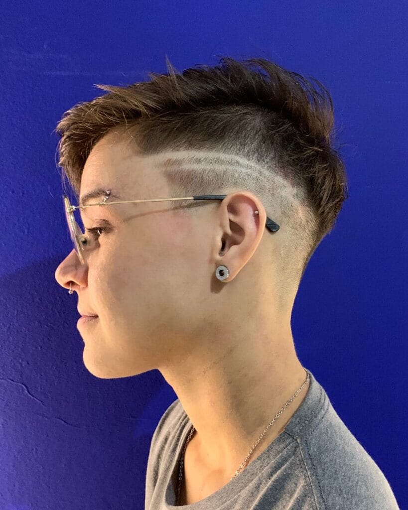 A woman with a sharp undercut tomboy haircut featuring a faded side, shaved line detail, and longer textured brown top.