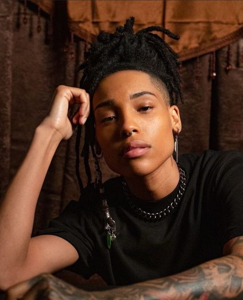 A woman with an undercut tomboy hairstyle featuring shaved sides and short locs styled upward on top.