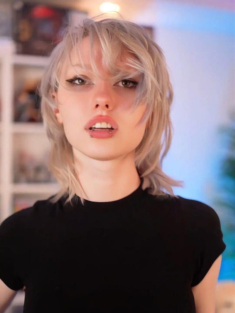 A young woman with a shaggy blonde tomboy mullet haircut featuring choppy layers and a wispy fringe.