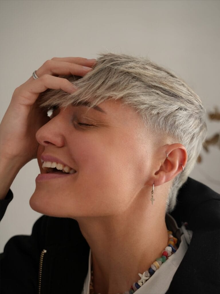 A young woman with a shaggy blonde tomboy mullet haircut featuring choppy layers and a wispy fringe.