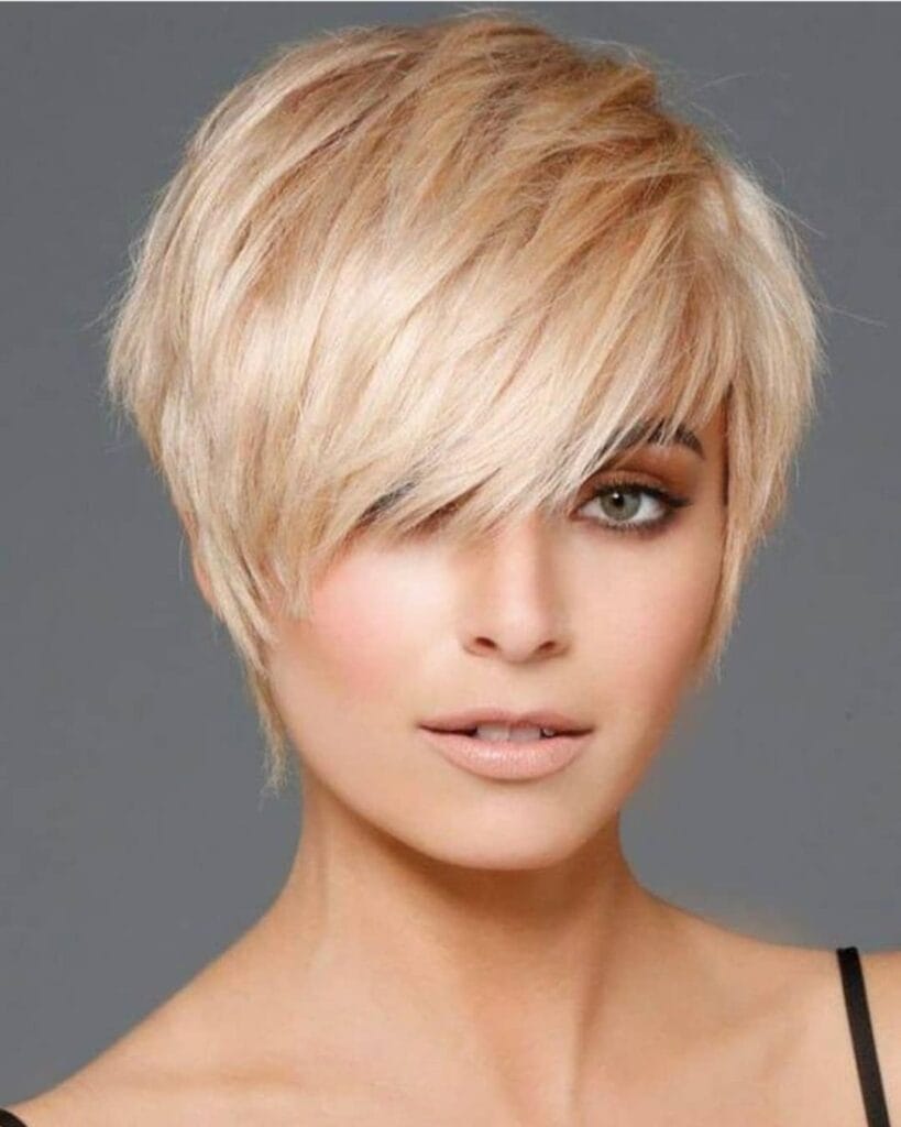 A short blonde pixie haircut with layered texture and a long side-swept fringe covering part of the forehead.