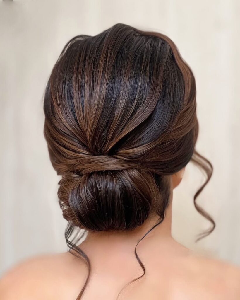 Sleek low twisted bun with smooth volume and soft curled face framing strands.