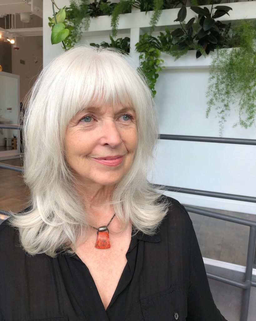 Long white gray layered hair with full straight bangs and soft feathered ends.