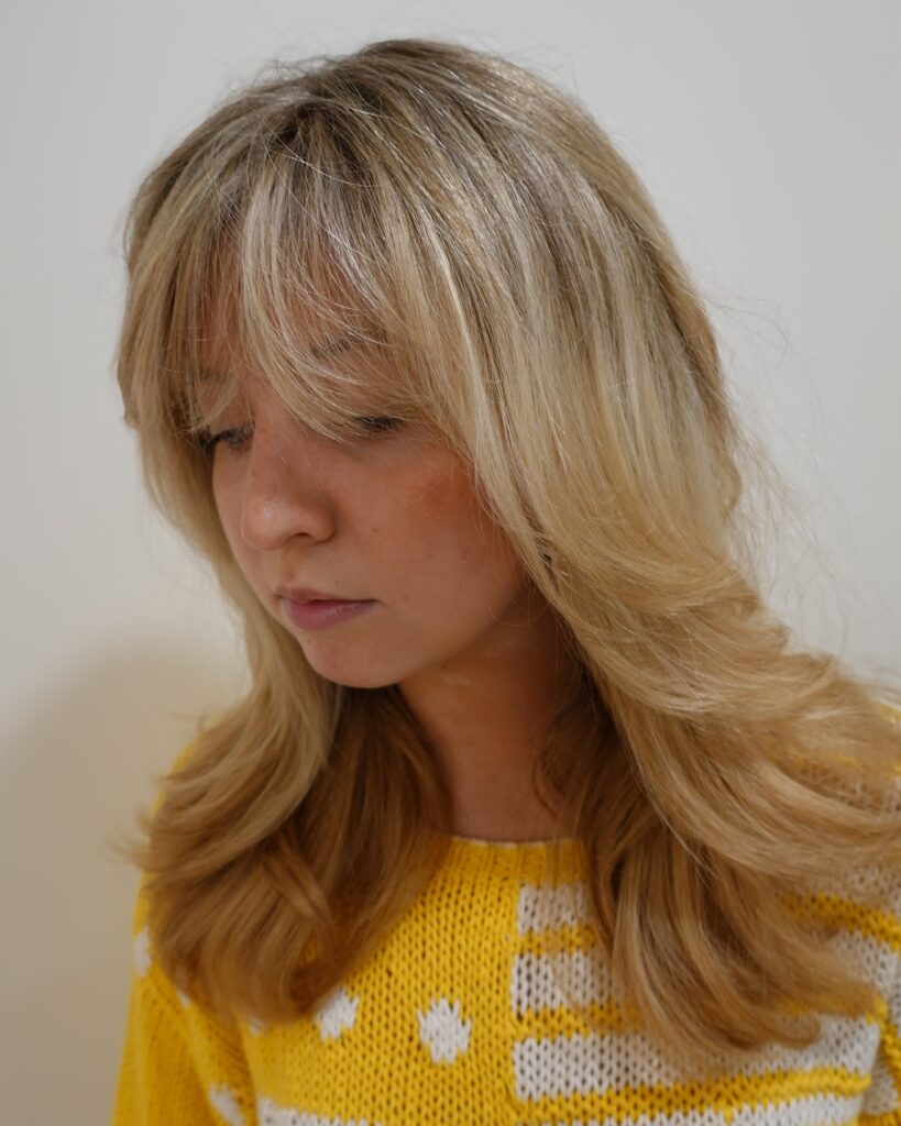 Medium length blonde layered hair with full feathered bangs and soft flipped ends.