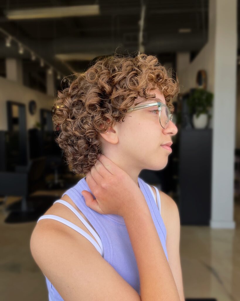 A woman with a short curly crop and soft fringe in light brown tones wearing glasses indoors.