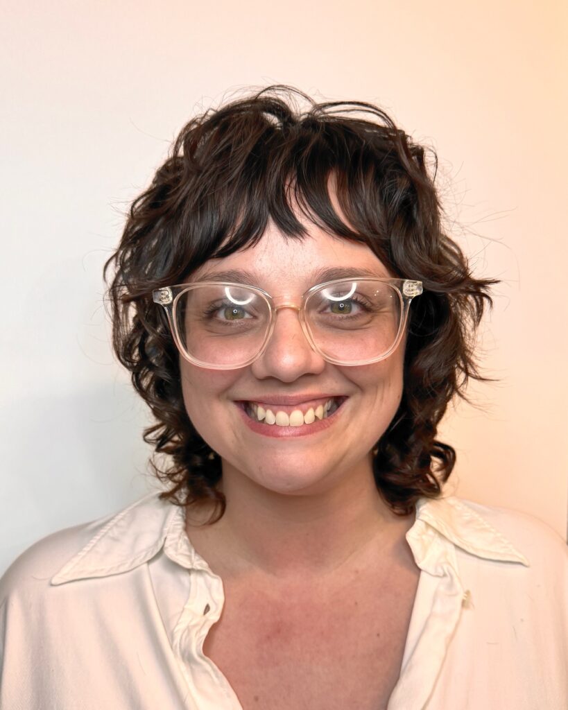 A woman with a soft layered curly bob and choppy bangs in dark brown tones wearing glasses.