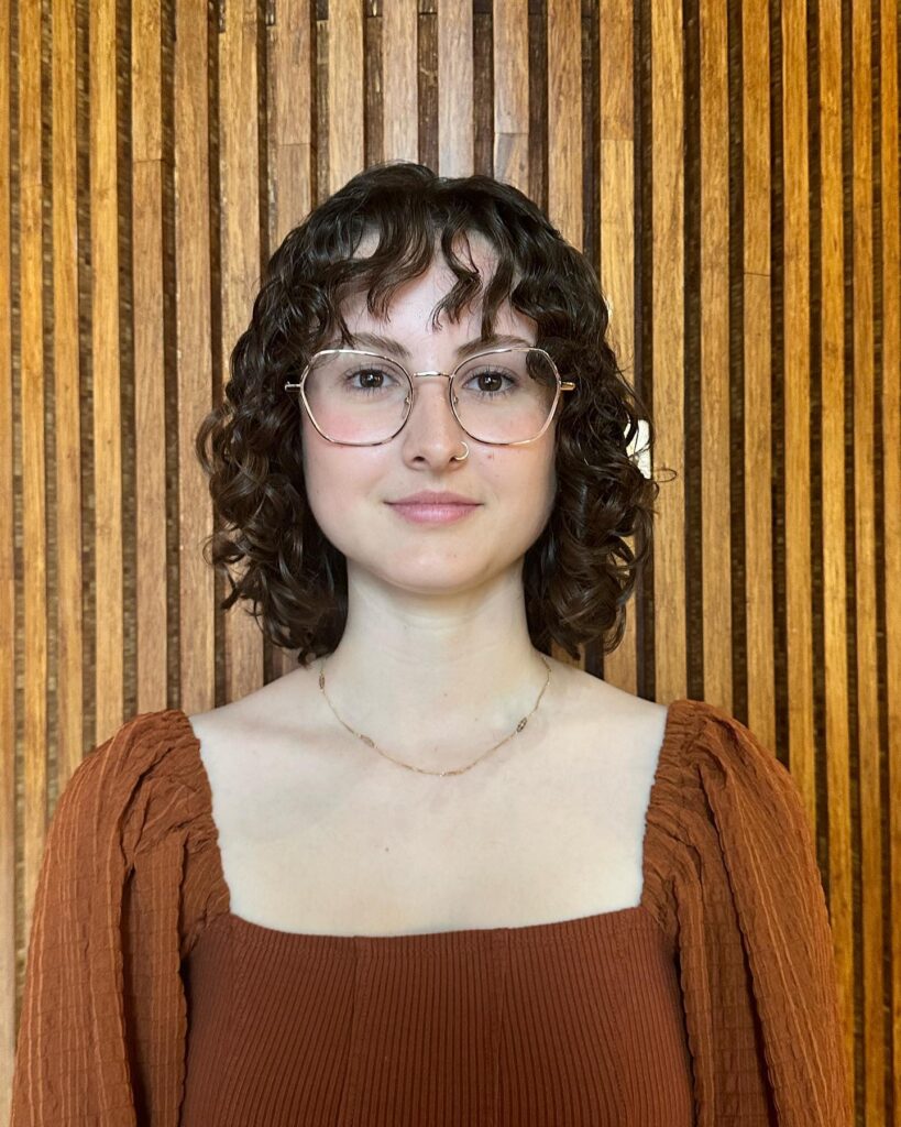 A woman with a natural curly bob and light fringe in dark brown tones standing in front of a wooden wall.