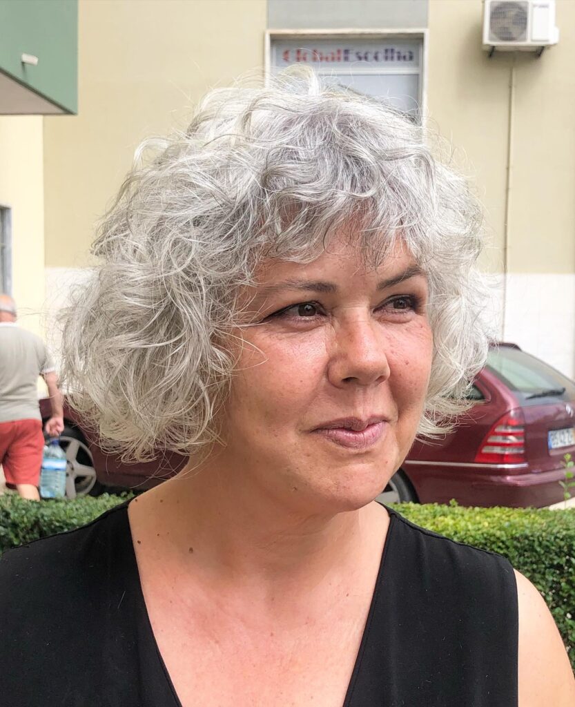 A woman with a voluminous silver curly shag and wispy bangs outdoors.