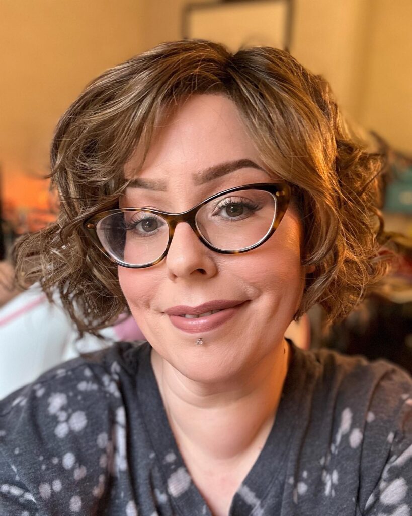 A woman with a soft curly bob and wispy bangs wearing glasses indoors.