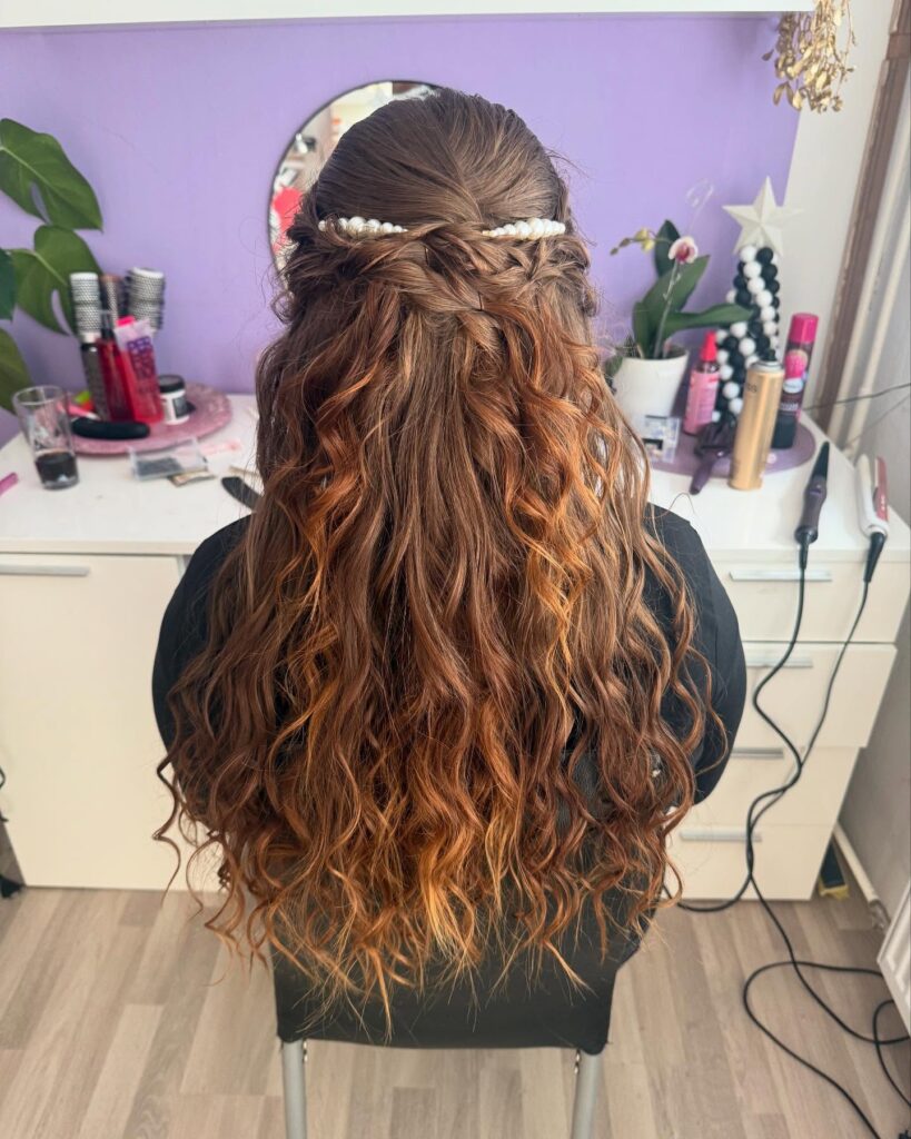 Long auburn curly hair styled in a half up braided crown with a pearl accessory across the back.