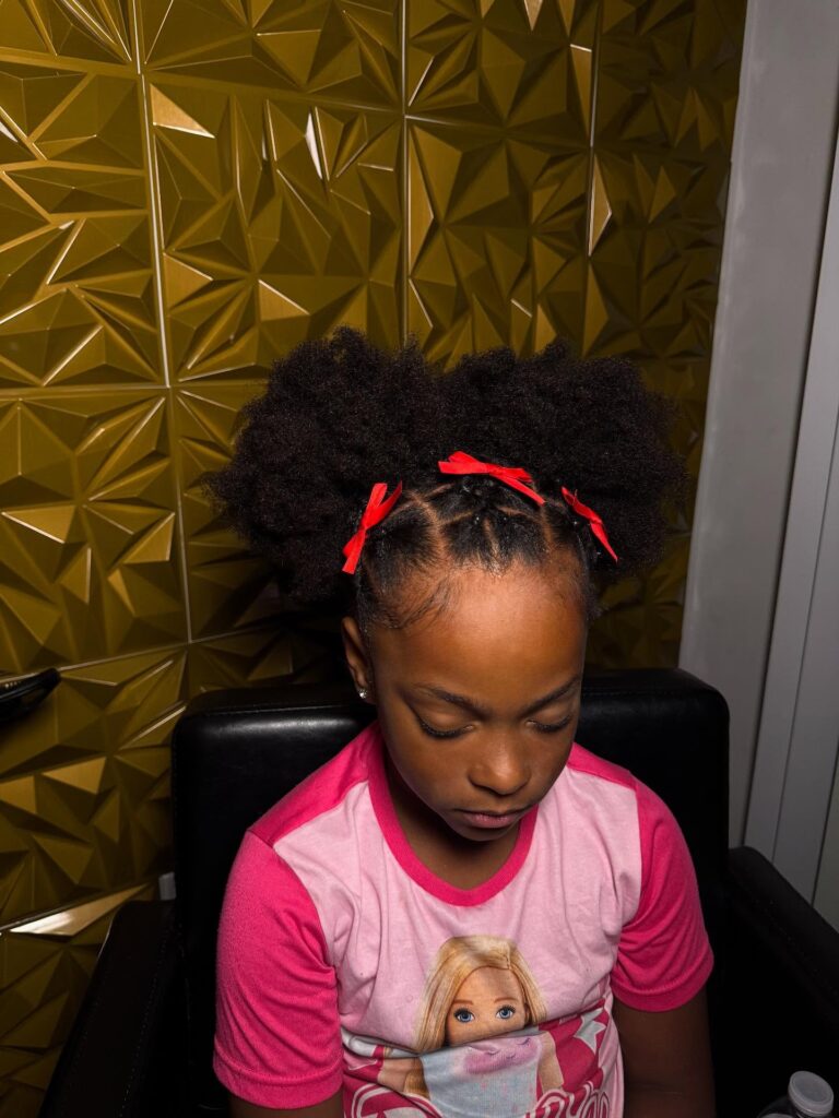 Natural hair styled into two puffs with front rubber band sections and red bow accessories.