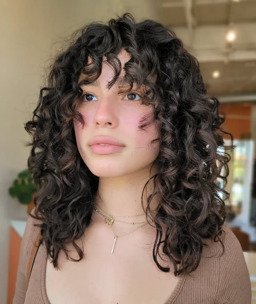 A woman with a voluminous curly lob and soft wispy bangs, featuring loose defined curls and natural texture.