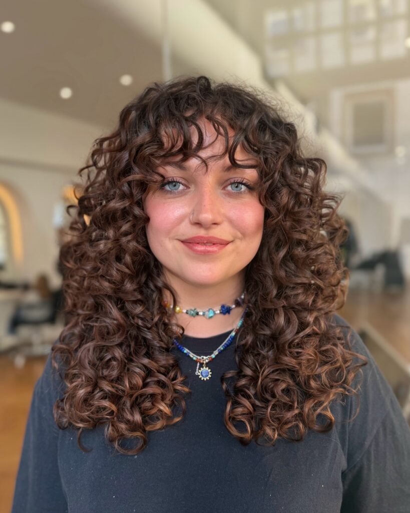 A woman with a defined curly bob and short micro bangs, showcasing tight ringlets and a rounded shape.