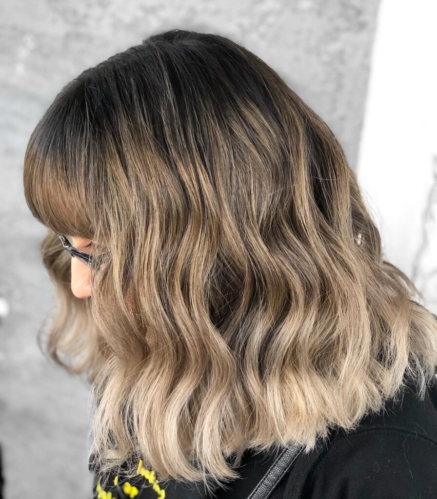 A woman with a soft layered lob and curtain bangs, featuring subtle waves and a natural brunette to blonde blend.