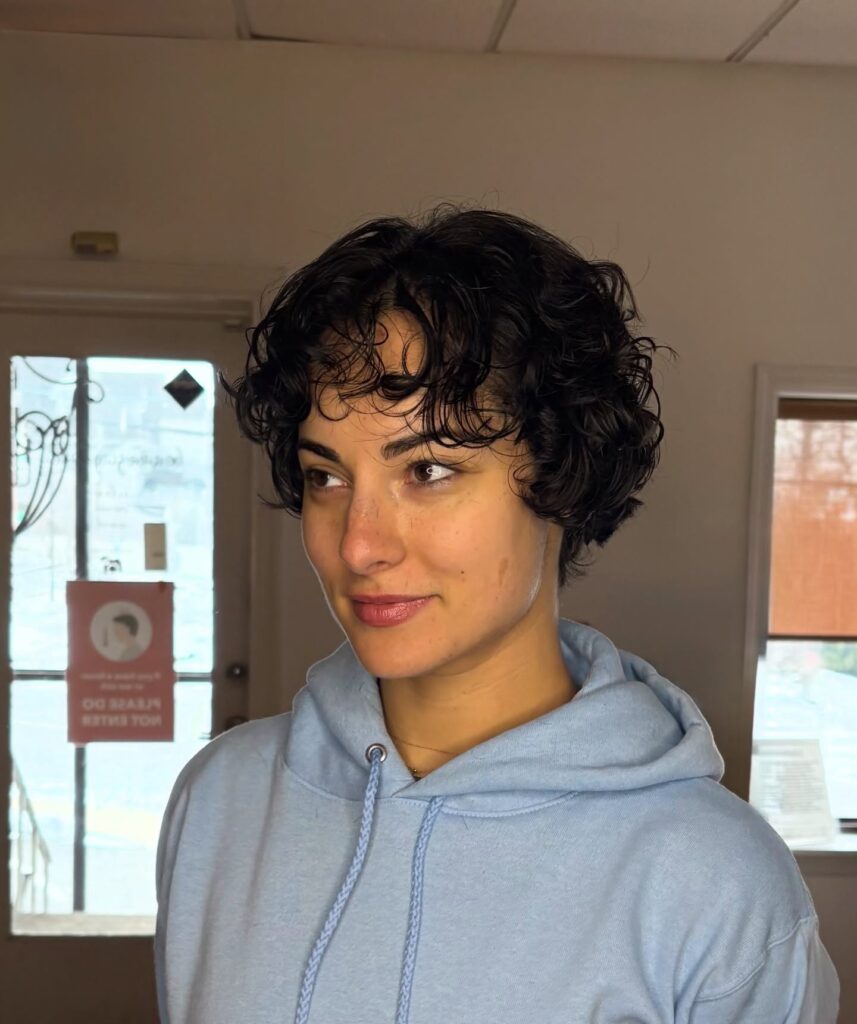 A woman with a short curly pixie haircut and soft wispy bangs in a dark brunette tone.