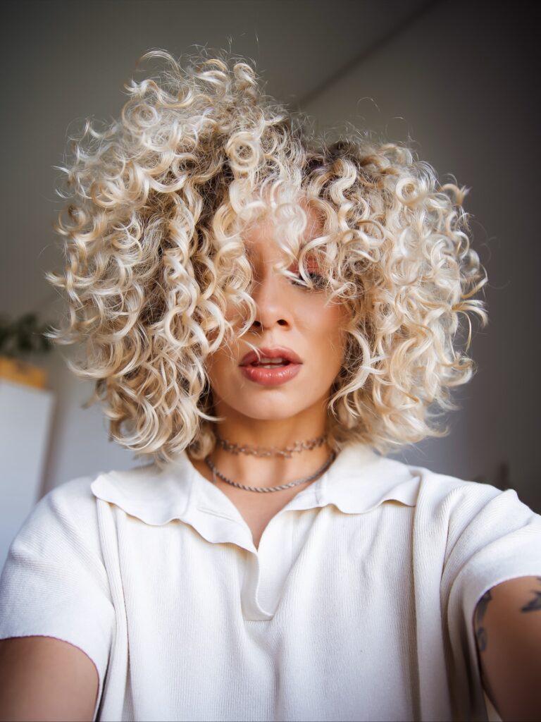 A woman with a short platinum blonde curly bob and full voluminous bangs covering the forehead.