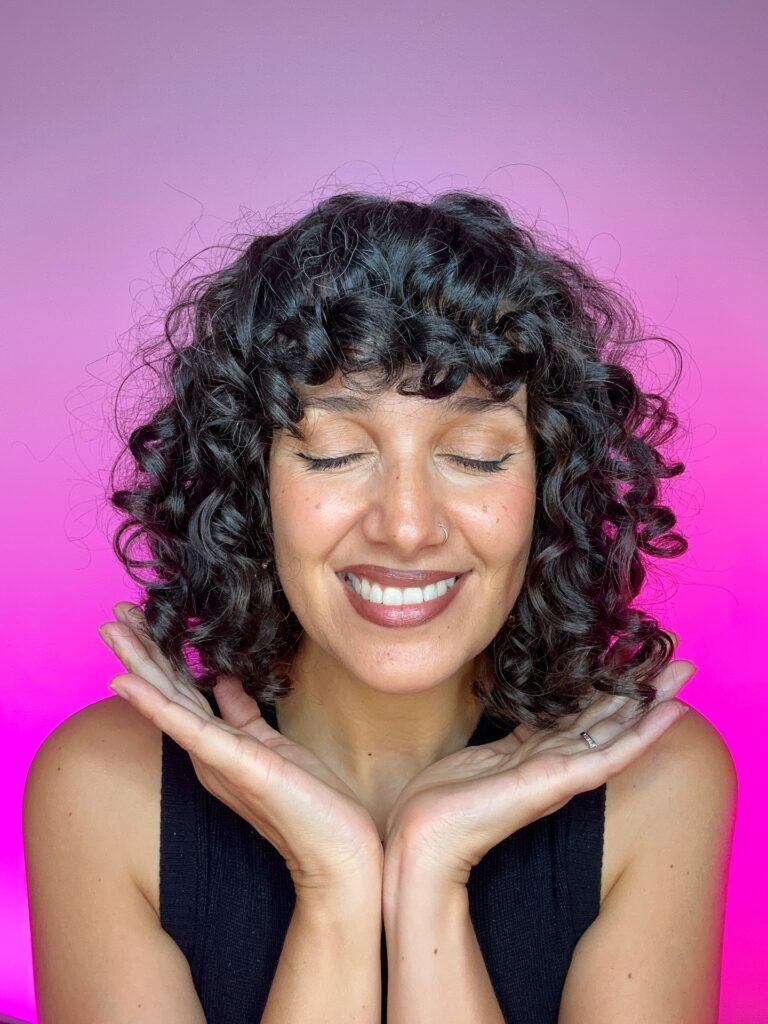 A woman with a short rounded curly bob and full bangs, featuring defined dark brown curls framing her face.