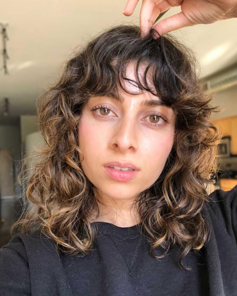 A woman with a tousled curly shag haircut and textured bangs, featuring loose curls and natural volume.