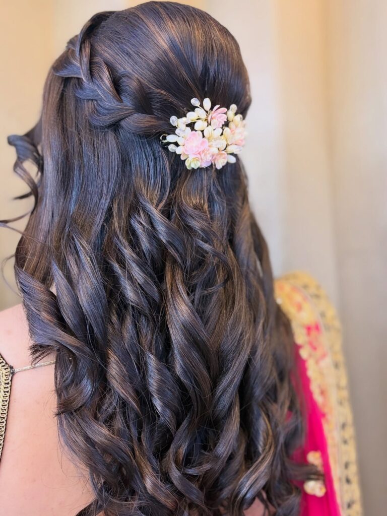 Long dark curly hair styled half up with defined curls and a floral hair accessory at the back.