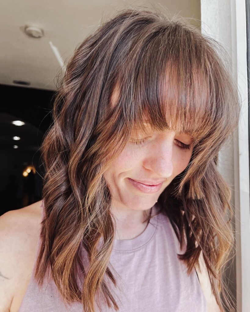 A textured shoulder length lob with soft waves and full blended bangs in light brown tones.