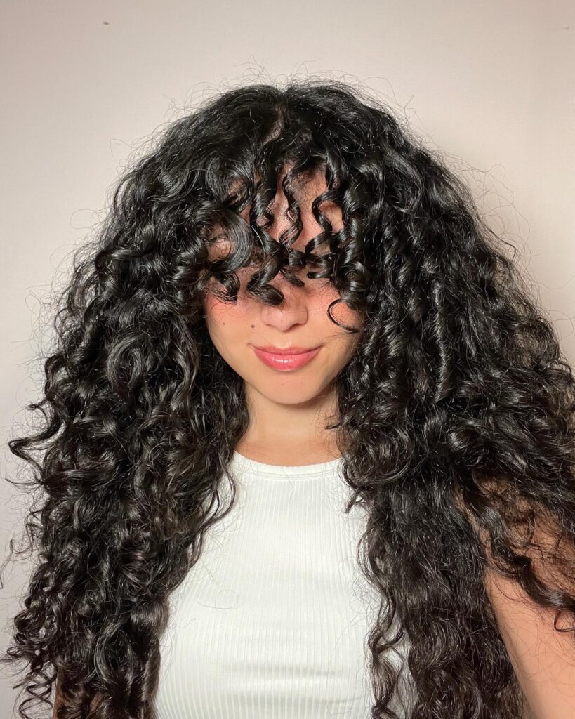 Long dark curly hair with full volume and a soft curly fringe covering the forehead.