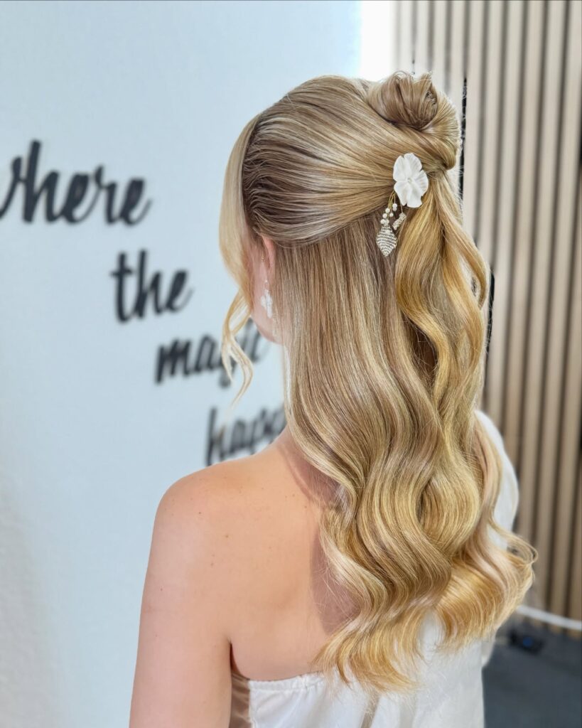 Long blonde wavy hair styled in a half up bun with a floral clip and soft waves.
