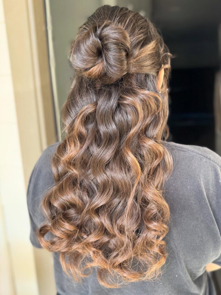 Long curly brunette hair with caramel highlights styled in a twisted half up look with flowing curls.