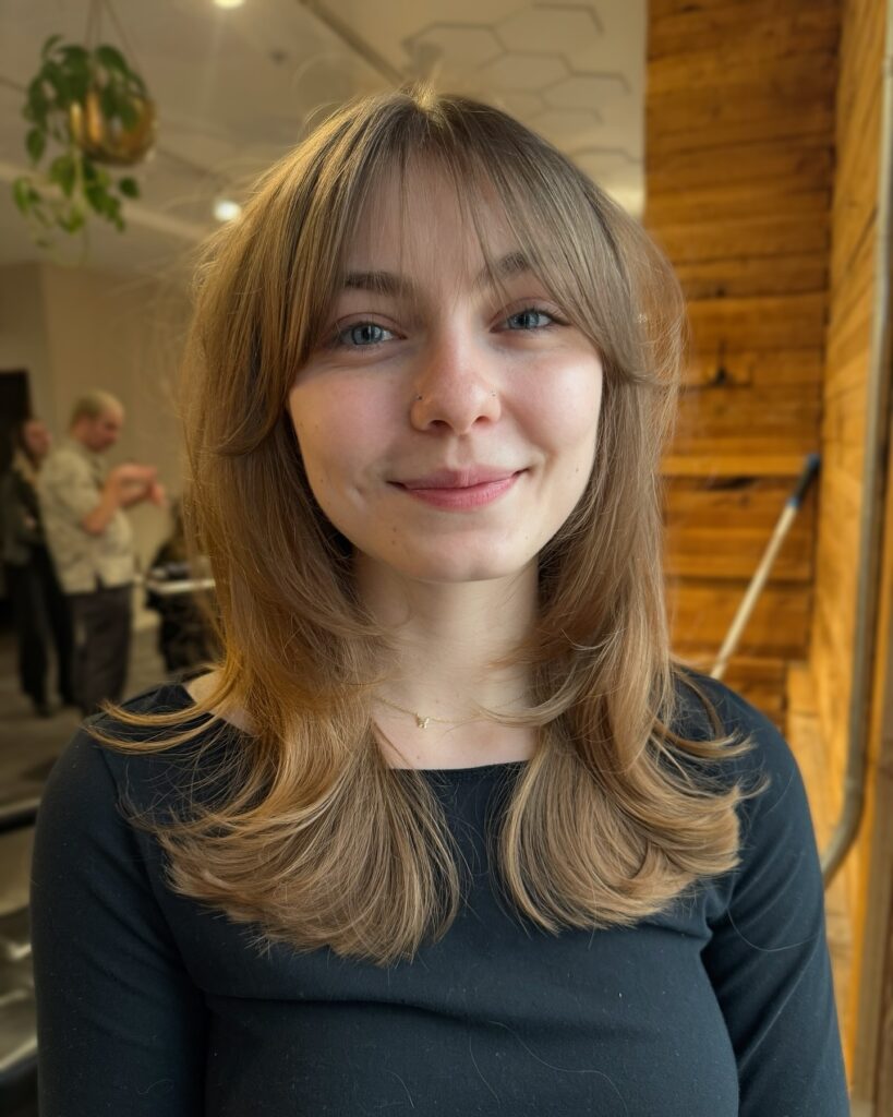 A woman with mid length feathered hair and soft wispy bangs in a natural blonde tone with light movement.