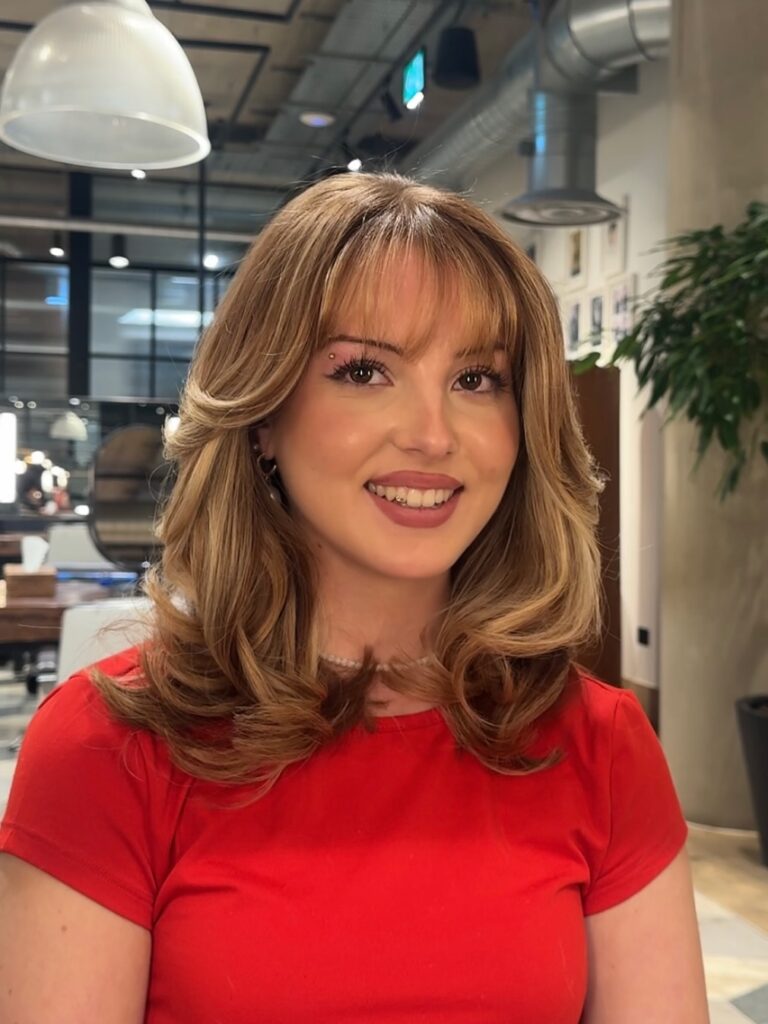 A woman with a shoulder length layered lob and soft wispy bangs styled with gentle volume and warm brown tones.