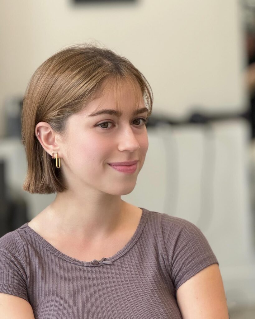 A woman with a sleek short bob haircut and subtle wispy bangs in a natural light brown shade.