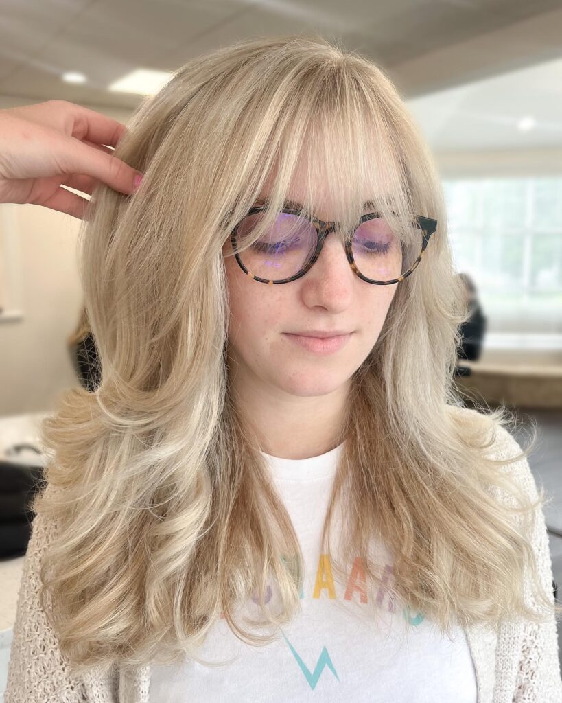A woman with long voluminous blonde hair and full wispy bangs styled with soft layers.