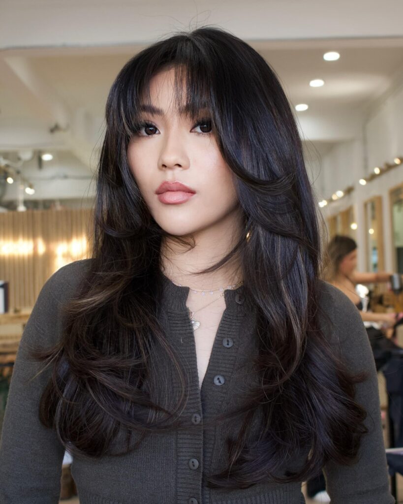 A woman with long layered dark hair and soft wispy bangs styled with smooth volume.