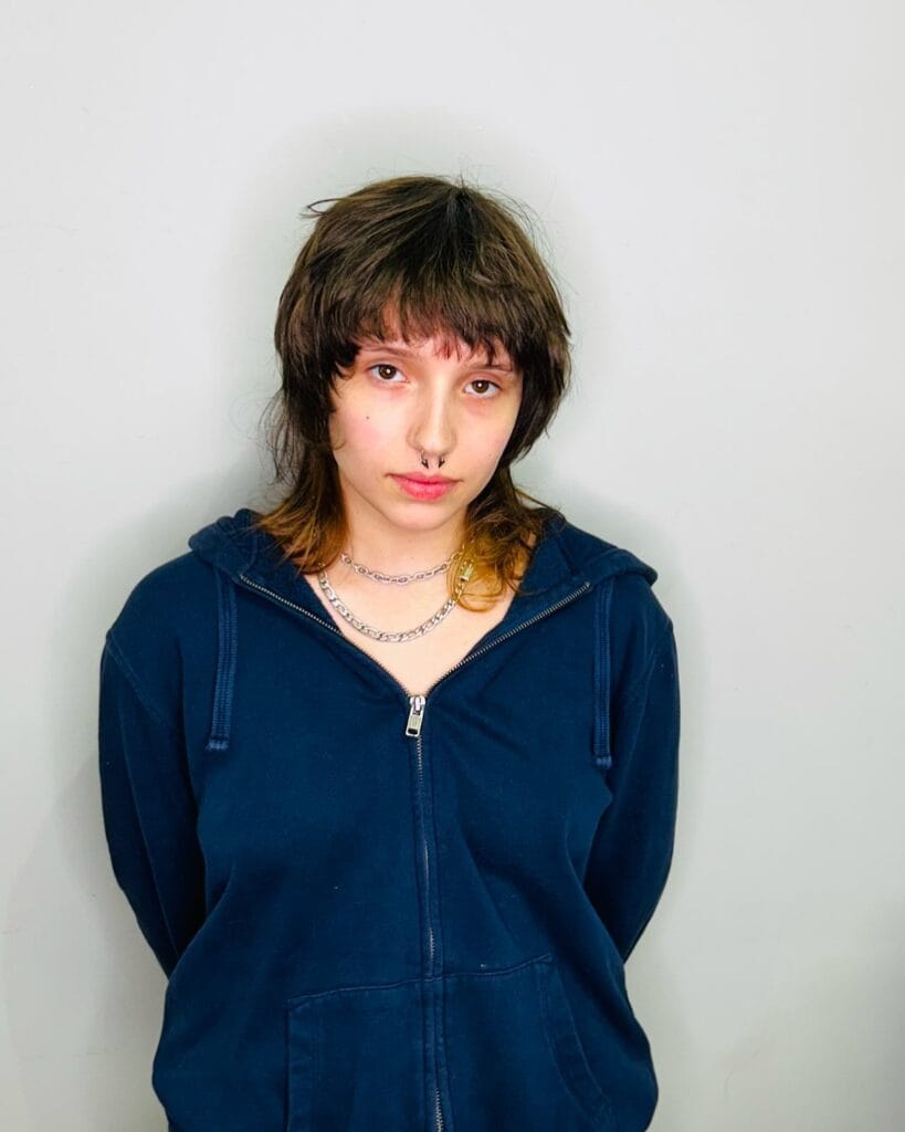 A woman with a short to medium shaggy tomboy haircut featuring soft layers, a light fringe, and natural brown hair with subtle lighter ends.