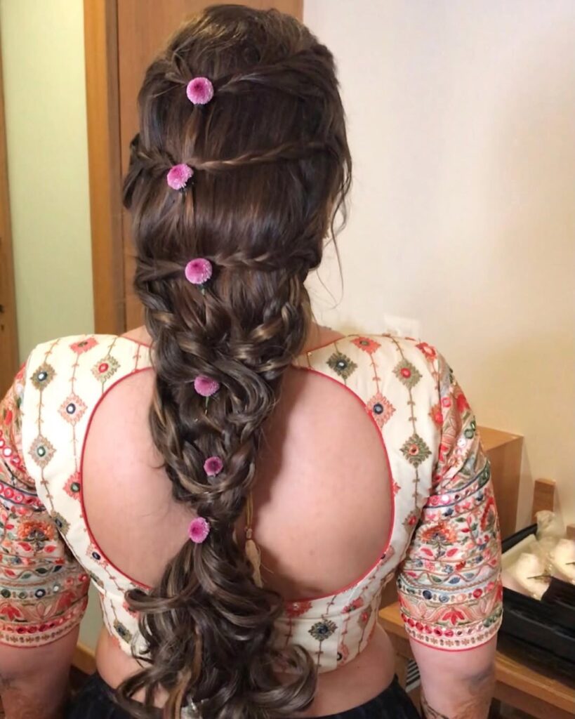 Long braided ponytail with loose curls and small pink floral accents placed along the braid.